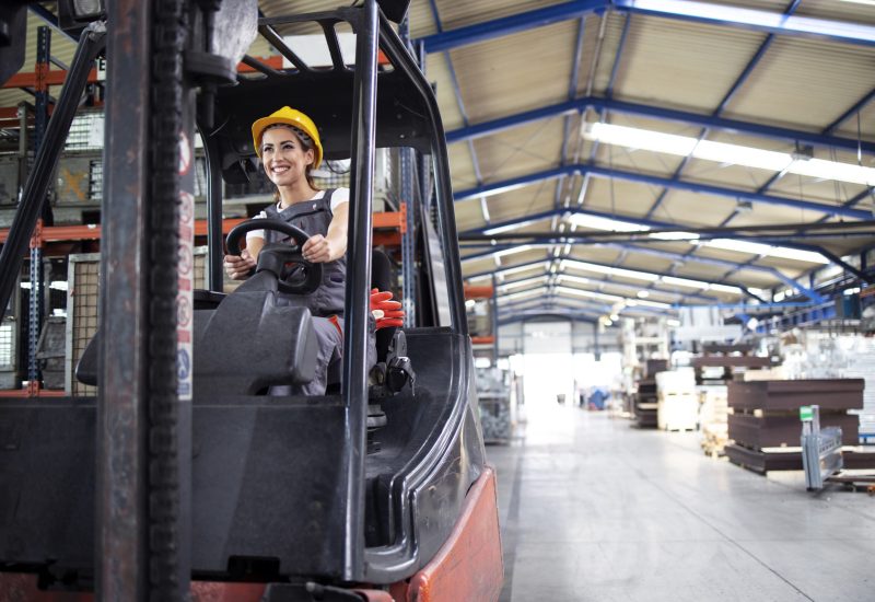 Professional female industrial driver operating forklift machine in factory's hall.
